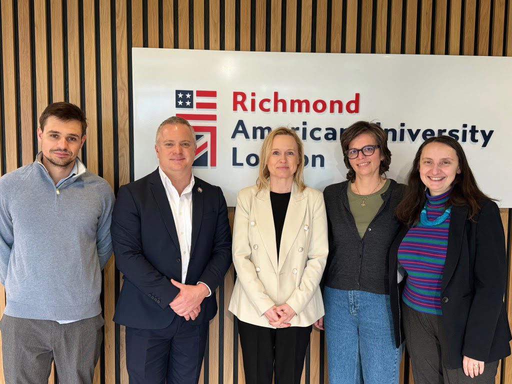 Five people are standing in front of a sign reading "RichmondAmerican University London," smiling casually for a photo in a wood-paneled room.