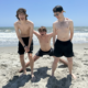 Three joyful young people are standing on a sandy beach with the ocean behind them. They are smiling and posing playfully under the sun.