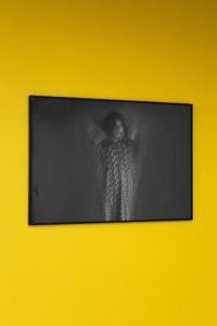 A black-and-white photograph of a person is displayed on a wall, featuring blurred movement against a vivid yellow background. The image has an abstract feel.