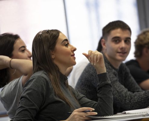 The image shows a group of young adults seated in a classroom, with one person gesturing and appearing engaged in a discussion. They seem focused and attentive.