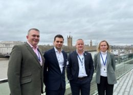 Four people stand on a balcony with the River Thames and Westminster, including the Houses of Parliament, in the background on an overcast day.