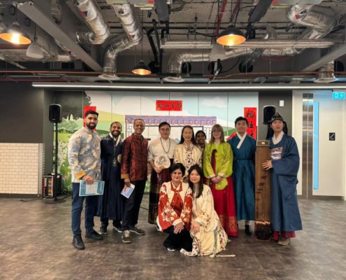 A diverse group of people pose together indoors. They wear traditional clothing, likely representing different cultures, against a modern, industrial background.