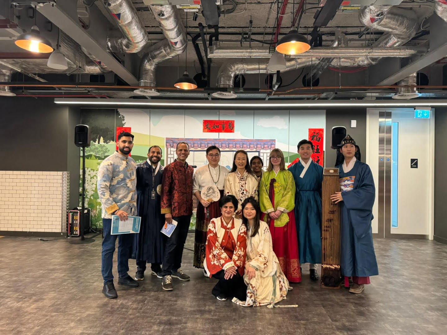 A diverse group of people pose together indoors. They wear traditional clothing, likely representing different cultures, against a modern, industrial background.
