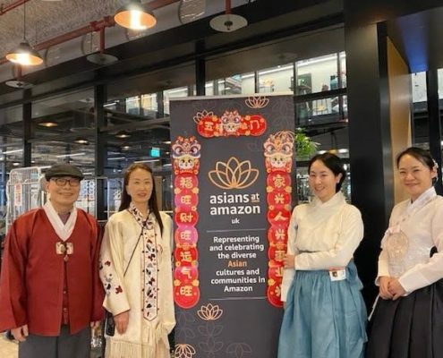 Four people stand next to a banner reading "asians at amazon uk," representing Asian cultures within the company. The interior setting appears modern.
