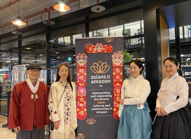 Four people stand next to a banner reading "asians at amazon uk," representing Asian cultures within the company. The interior setting appears modern.
