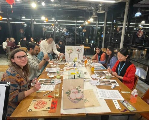 A group of people is attending a brush painting workshop. They're seated at tables with painting supplies, focused on creating artwork.
