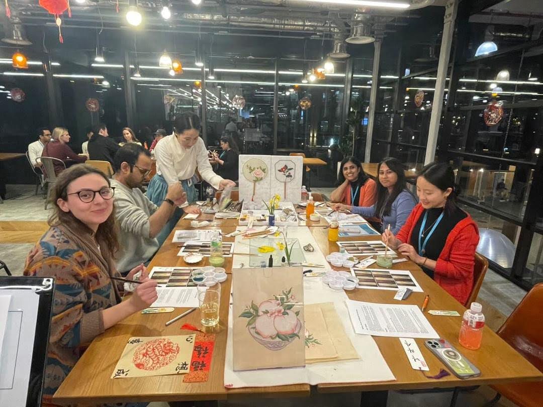 A group of people is attending a brush painting workshop. They're seated at tables with painting supplies, focused on creating artwork.