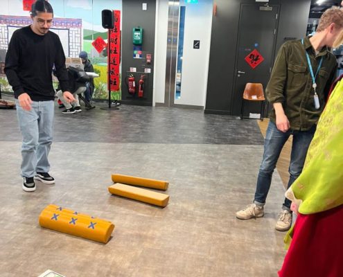 Two people are standing in a modern indoor space with exposed ceiling pipes. There's a board game on the floor and colorful stools nearby.