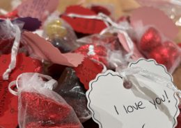 The image shows a collection of red heart-shaped chocolates with personalized notes attached, expressing love and affection, possibly for Valentine's Day.